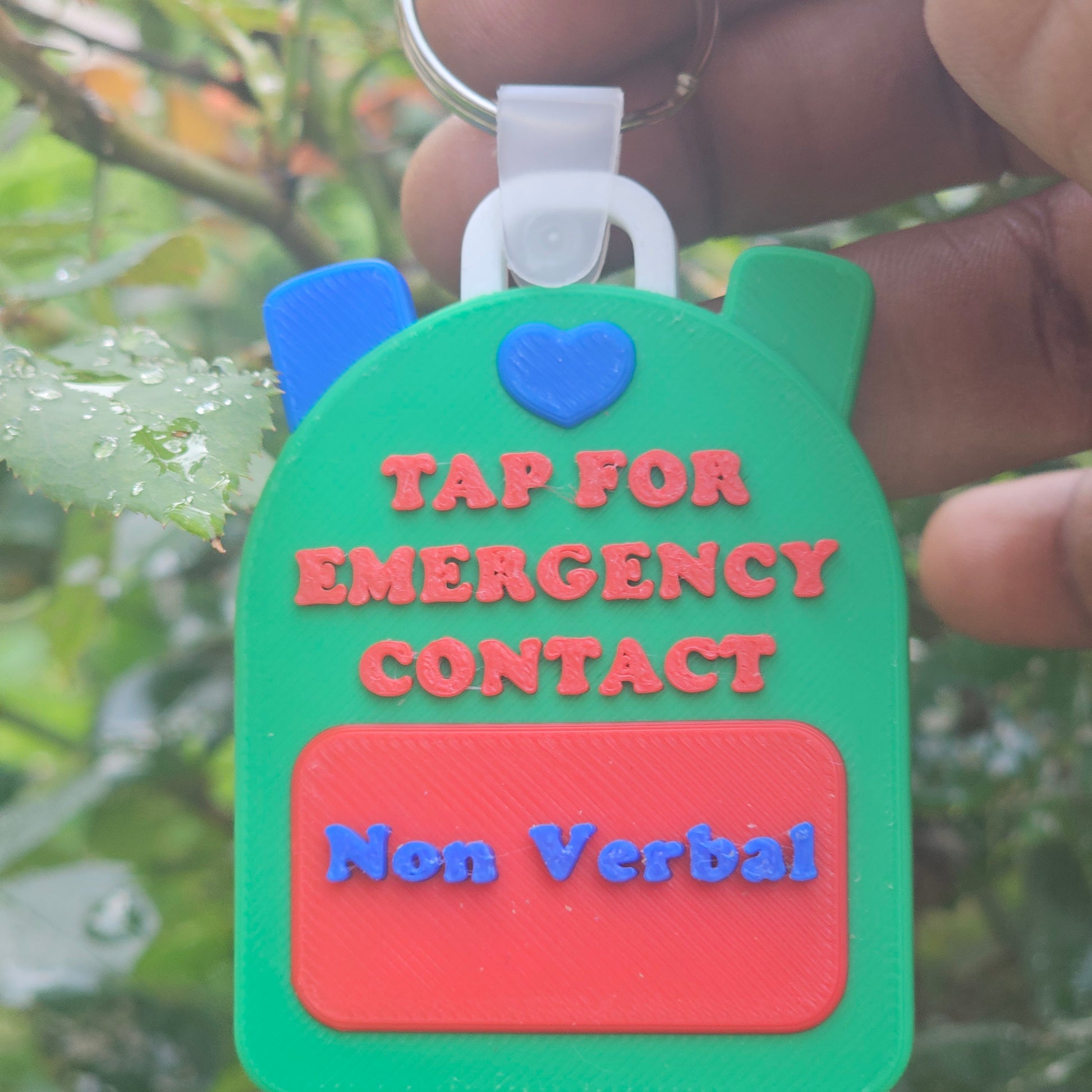 Green keychain with text 'Tap for Emergency Contact Non Verbal' held by a hand against a blurred natural background.