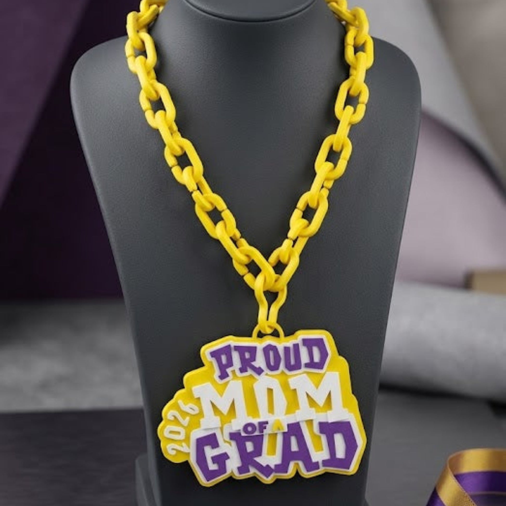 Yellow 'Proud Mom of Grad' necklace on a black stand with a purple box labeled 'Graduation Keepsakes Collection'.