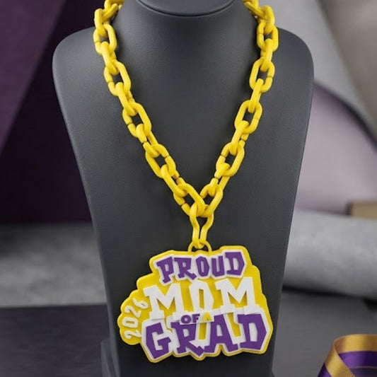 Yellow 'Proud Mom of Grad' necklace on a black stand with a purple box labeled 'Graduation Keepsakes Collection'.